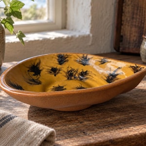 May include: A shallow, triangular ceramic bowl with a mustard yellow interior featuring scattered black brushstroke patterns. The exterior of the bowl has a textured, earthy brown finish, and the overall shape is asymmetrical with rounded corners.