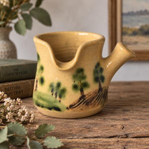 Peut inclure: Un pot à lait en céramique jaune avec une anse latérale et un bec verseur. L'extérieur présente un motif paysager peint à la main avec des arbres verts et des détails de sol bruns. Le récipient a une texture mouchetée et une large ouverture.