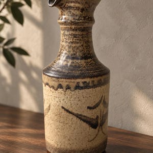 May include: A tall ceramic pitcher with a narrow neck and a flared spout. The upper portion features a dark brown, textured glaze, while the lower body is a speckled cream color with dark brown abstract brushstroke patterns around the base.