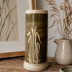 Pode incluir: Um vaso cilíndrico de cerâmica com um esmalte verde-oliva escuro e um acabamento texturizado de pinceladas. A superfície apresenta um motivo esculpido de talos de trigo em cor clara. O vaso tem uma borda e base cor de creme sem esmalte.