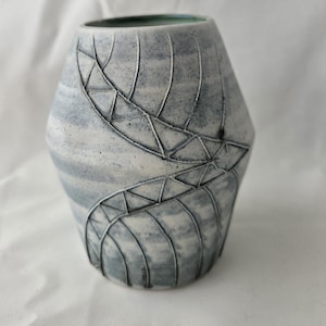 May include: A ceramic vase with a tapered cylindrical shape, featuring a matte white and grey mottled finish. The surface is decorated with deep, dark grey incised lines forming a continuous, winding leaf-like pattern that wraps around the body.