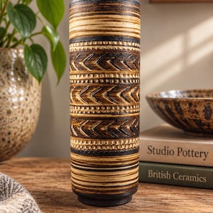 May include: A cylindrical ceramic vase featuring a dark brown and cream colour palette. The exterior is decorated with horizontal bands of incised chevron patterns and fine linear grooves, creating a textured surface throughout the entire vessel.
