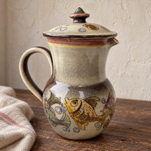 Può includere: Una brocca in ceramica con coperchio, caratterizzata da una smaltatura sfumata beige e marrone. Il corpo è decorato con motivi di pesci dipinti a mano in giallo e multicolore e piccoli cerchi blu che rappresentano bolle. Manico e beccuccio.