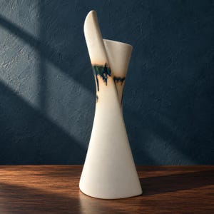 May include: A tall, off-white ceramic vase with a flared base and a twisted, asymmetrical rim. The upper section features a decorative drip glaze in shades of dark blue and green that cascades down from the opening onto the smooth, matte body.