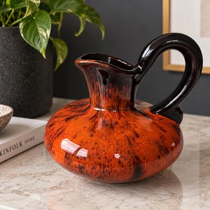 May include: A ceramic pitcher featuring a wide, rounded base and a glossy finish with a vibrant orange and black mottled glaze. The piece includes a curved, solid black handle and a flared spout, showcasing a smooth, reflective surface texture.