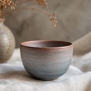 May include: A small, round ceramic bowl featuring a gradient finish that transitions from a warm, reddish-brown tone at the rim to a cool, muted blue-grey colour at the base. The surface has a matte, textured appearance with visible horizontal striations.