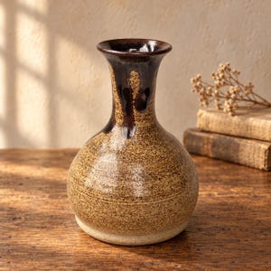 May include: A ceramic vase with a bulbous body and a flared rim. The surface features a speckled tan and brown finish, with a dark brown glaze dripping from the rim down the neck. The base is a lighter, solid cream colour with visible texture.
