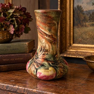 May include: A ceramic vase featuring a relief design of leaves and fruit in shades of green, brown, and red. The vase has a flared rim and a bulbous base with a textured, multi-colored glaze finish that covers the entire exterior surface.