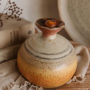 May include: A round ceramic vase with a narrow neck and a flared rim. The body features a gradient glaze transitioning from a creamy off-white at the top to a warm, textured golden-orange at the base. A small, smooth wooden sphere rests inside the opening.