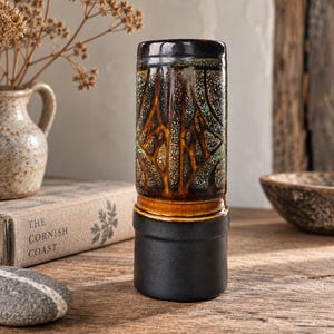 May include: A cylindrical ceramic vase featuring a glossy brown and green mottled glaze on the upper section with a textured, dark brown, tree-like pattern, transitioning to a smooth, matte black base with a distinct horizontal ridge.