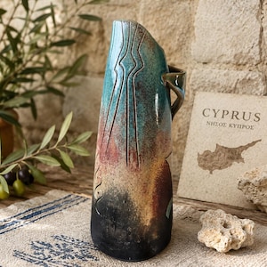 May include: A tall, tapered ceramic vase featuring a gradient glaze that transitions from a deep black base to a warm amber, then a vibrant turquoise top. The surface is decorated with etched geometric lines and has a single side handle.