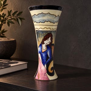 May include: A tall, flared ceramic vase with a cream-colored base and a dark blue rim. The exterior features a hand-painted illustration of a person with brown hair wearing a blue top and a pink garment, set against a stylized, abstract background.