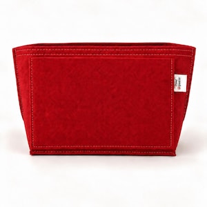 Puede incluir: Un organizador de bolso de fieltro rojo brillante con forma rectangular, que presenta un bolsillo frontal grande y costuras visibles a lo largo de los bordes. Una pequeña etiqueta de tela blanca en el lado derecho muestra el texto Trend Organizer.