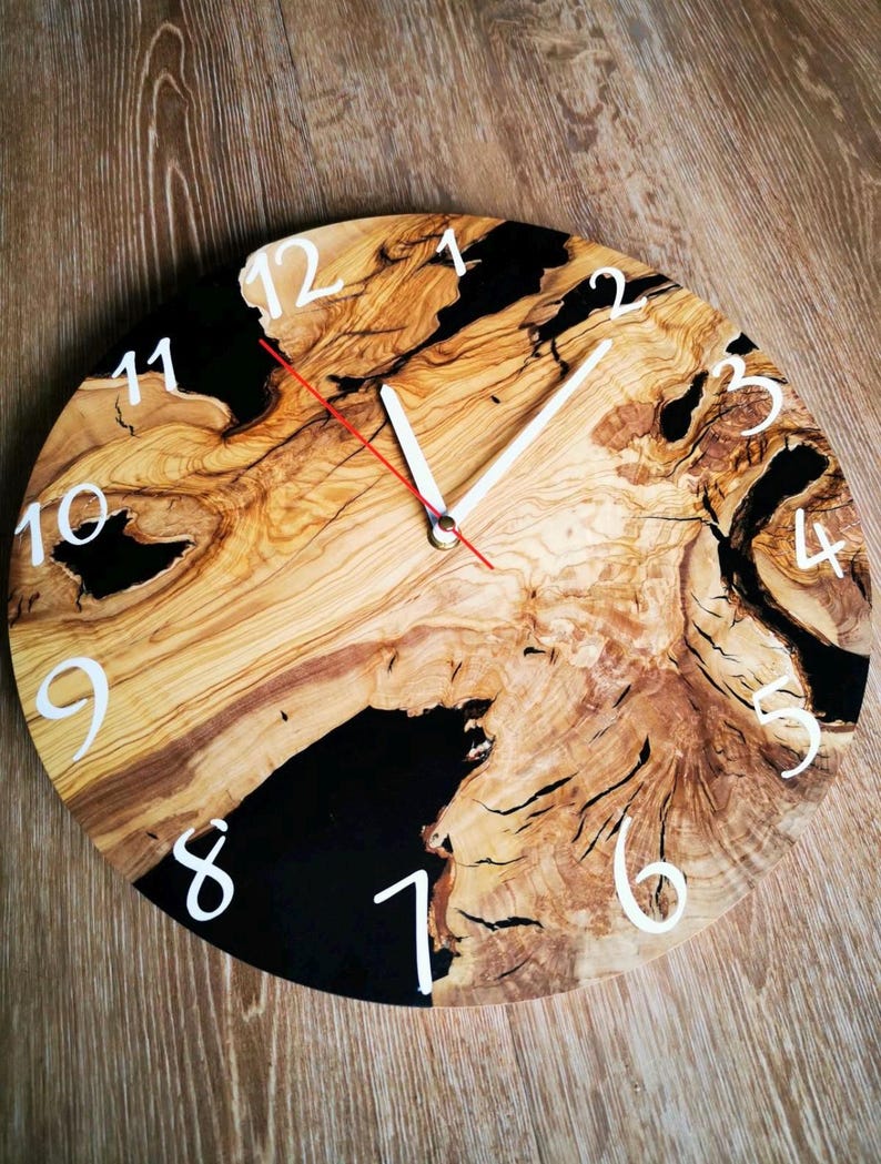 Op de afbeelding: Een ronde houten klok met zwarte epoxyhars accenten. De wijzerplaat van de klok heeft witte Romeinse cijfers en witte wijzers.