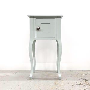 May include: A pale blue bedside cabinet with a single door and a silver knob. The table has curved legs and a rectangular top. This small cabinet is a piece of bedroom furniture.