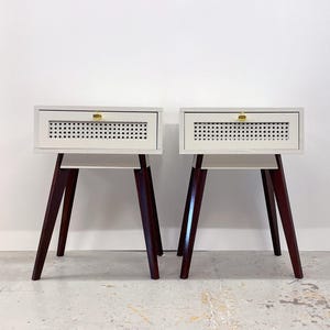 May include: A pair of matching mid-century modern bedside tables. Each has a light-coloured rectangular drawer with a perforated front and a gold-coloured knob. The tables have dark brown, angled legs and a lower shelf.
