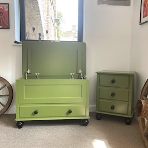 May include: A green storage chest with two drawers and a matching three-drawer bedside table. The chest's lid is open, revealing the interior. A wooden cartwheel and framed art pieces complete the room's decor.
