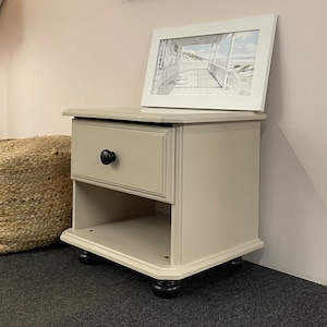 May include: A beige bedside table with a single drawer and a lower shelf. The drawer has a black knob. A framed picture of a beach scene rests on top. The table is on black feet.