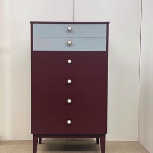 May include: A tall, mid-century modern style chest of drawers with six drawers. The top two drawers are light blue, while the bottom four are a deep burgundy. Each drawer has a white, round knob. The chest stands on four tapered legs.