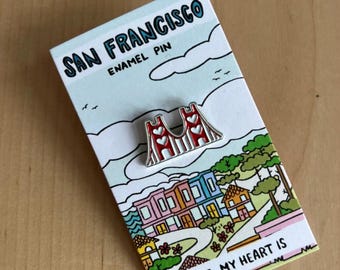 May include: A red and white enamel pin shaped like the Golden Gate Bridge, attached to a rectangular display card featuring a colorful illustration of San Francisco houses and the text SAN FRANCISCO ENAMEL PIN and WHERE MY HEART IS.