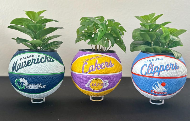 May include: Three basketball-themed planters, each with a different team logo: Dallas Mavericks, Los Angeles Lakers, and San Diego Clippers. Each planter contains a vibrant green plant, creating a unique decorative item.