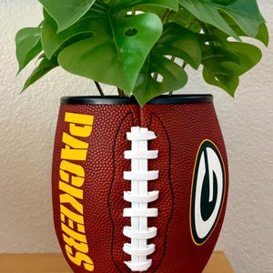 Pode incluir: Um vaso texturizado castanho em forma de bola de futebol americano com a palavra PACKERS em letras amarelas e um logótipo G verde e amarelo. O vaso tem atacadores brancos ao centro e contém uma planta verde com várias folhas.