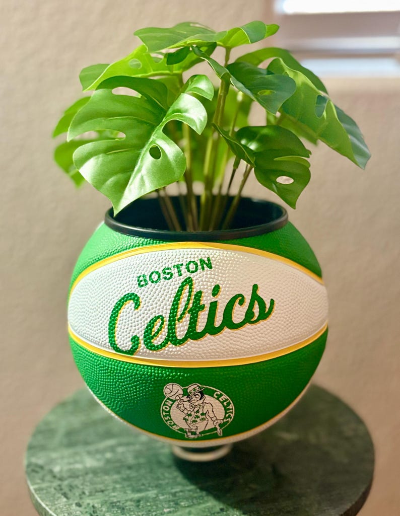 May include: A green and white basketball planter with the Boston Celtics logo and text. It contains a green leafy plant. The planter is on a green marble table.