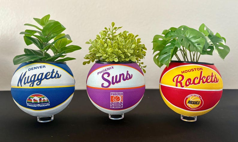 May include: Three basketball-themed planters, each with an NBA team logo: Denver Nuggets, Phoenix Suns, and Houston Rockets. Each planter holds different greenery, creating a decorative display. The planters are supported by clear stands.