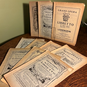 May include: A collection of vintage Grand Opera Libretto booklets. The covers are tan with black text and illustrations. Titles include "Lohengrin" and "Don Pasquale". The booklets are arranged on a wooden surface.