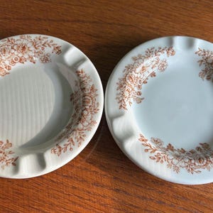 May include: Two vintage ashtrays with a floral design. The ashtrays are round with a light cream color and a brown floral pattern around the rim. Each ashtray has three notches for resting cigarettes. The ashtrays are made of ceramic.