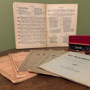 May include: A collection of vintage opera programmes, sheet music, and opera glasses in a red case. The items are arranged on a wooden surface. The sheet music is open, displaying musical notes.