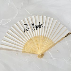 May include: A white and cream folding fan with the words "The Bride" in black script. The fan has a wooden handle and the name "Clarissa" is printed on the side. The fan is open and displayed on a white surface.