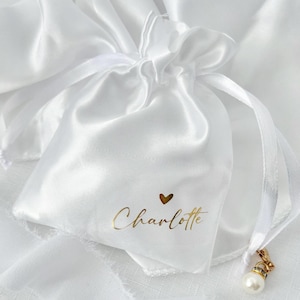 May include: A white satin drawstring pouch featuring the name Charlotte printed in gold script lettering with a small gold heart icon above the text. A single white pearl bead charm with a gold-toned metal clasp hangs from the ribbon tie.
