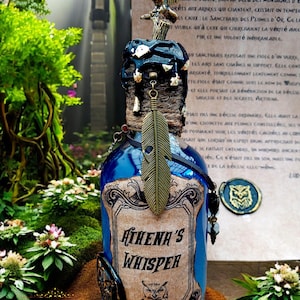Peut inclure: Une bouteille en verre bleue avec l'inscription "Athena's Whisper" et une illustration de hibou. La bouteille est ornée d'une plume décorative, d'un hibou en métal et d'un bouchon détaillé. Entourée de verdure.