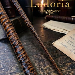 Puede incluir: Dos varitas de madera talladas a mano con un acabado marrón oscuro y una superficie texturizada y estriada. Las varitas presentan un diseño cónico con un mango grueso y una punta fina. La palabra Ludoria aparece en texto dorado arriba.