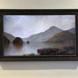 May include: A framed landscape painting depicting a serene lake scene with mountains in the background. The artwork features a dark, ornate frame and a muted color palette of blues, greens, and browns. The painting is mounted on a white wall.