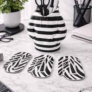 May include: A desk tidy in the shape of a hoodie, with black and white horizontal stripes, holding scissors and pens. Three matching zebra-striped coasters are in front. A watch and a laptop are also visible.