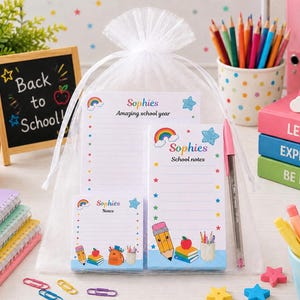 May include: A white organza drawstring bag containing three personalised stationery notepads with rainbow, star, and school supply graphics. The pads feature the name Sophies and text including Amazing school year and School notes. A pink pen is included.
