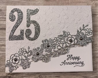 May include: A white greeting card featuring the number 25 in large silver glitter, a decorative silver glitter floral die-cut border with small dark beads, and the text Happy Anniversary printed in black script on the lower right corner.