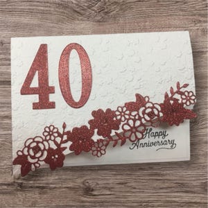 May include: White anniversary card with a textured surface, featuring a large, glittery red "40" and a decorative floral border. The words "Happy Anniversary" are printed in black.