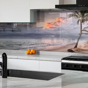 May include: A kitchen with white cabinets and a black range hood. The backsplash is a glass panel with a beach scene featuring palm trees, a sunset, and a sandy beach.