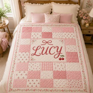 May include: A quilted bedspread featuring a patchwork design of pink and white fabrics with cherry, floral, and gingham patterns. The center panel displays the name Lucy in red cursive embroidery, accented by a red bow and two cherries.