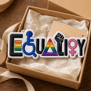 May include: An embroidered patch spelling the word EQUALITY, where the letter E features horizontal rainbow stripes, the Q is a blue wheelchair symbol, the A is a rainbow triangle, the L is a black raised fist, and the I is a pink female symbol.