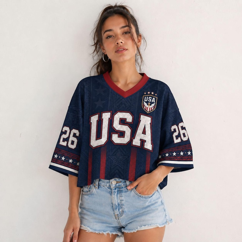 A young woman wearing a navy blue USA jersey with the number 26, paired with denim shorts, stands against a white background.