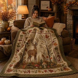 May include: A quilted throw blanket featuring a central illustration of a deer standing among trees, ferns, and mushrooms. The design is bordered by a pattern of autumn leaves in shades of orange, red, and green on a cream-colored background.