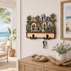 May include: A wall-mounted key holder featuring a three-dimensional Los Angeles cityscape with palm trees, a vintage car, a surfboard, and street signs. The wooden base includes two black metal double hooks, with a set of keys hanging from one.