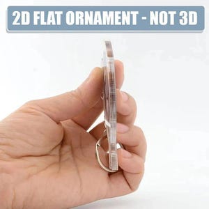 A hand holding a clear plastic ornament, which appears to be a 2D flat ornament.