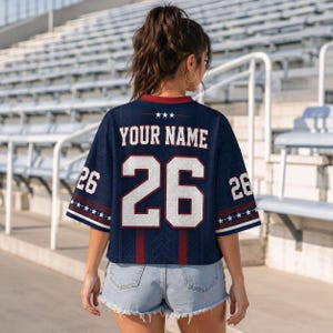 A woman wearing a navy blue jersey with the number 26 and the text "YOUR NAME" printed on the back, standing in front of a stadium seating area.