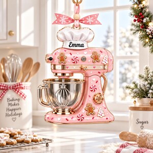 A pink and gold kitchen mixer with a chef's hat and a personalized name tag, surrounded by baking supplies and a festive holiday decoration.