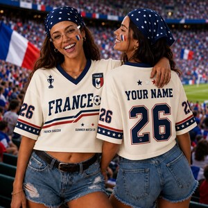 Two women wearing France soccer jerseys and bandanas are smiling and posing together in a stadium.